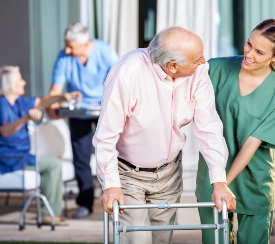 Happy female caretaker assisting senior man in using Zimmer frame at nursing home yard